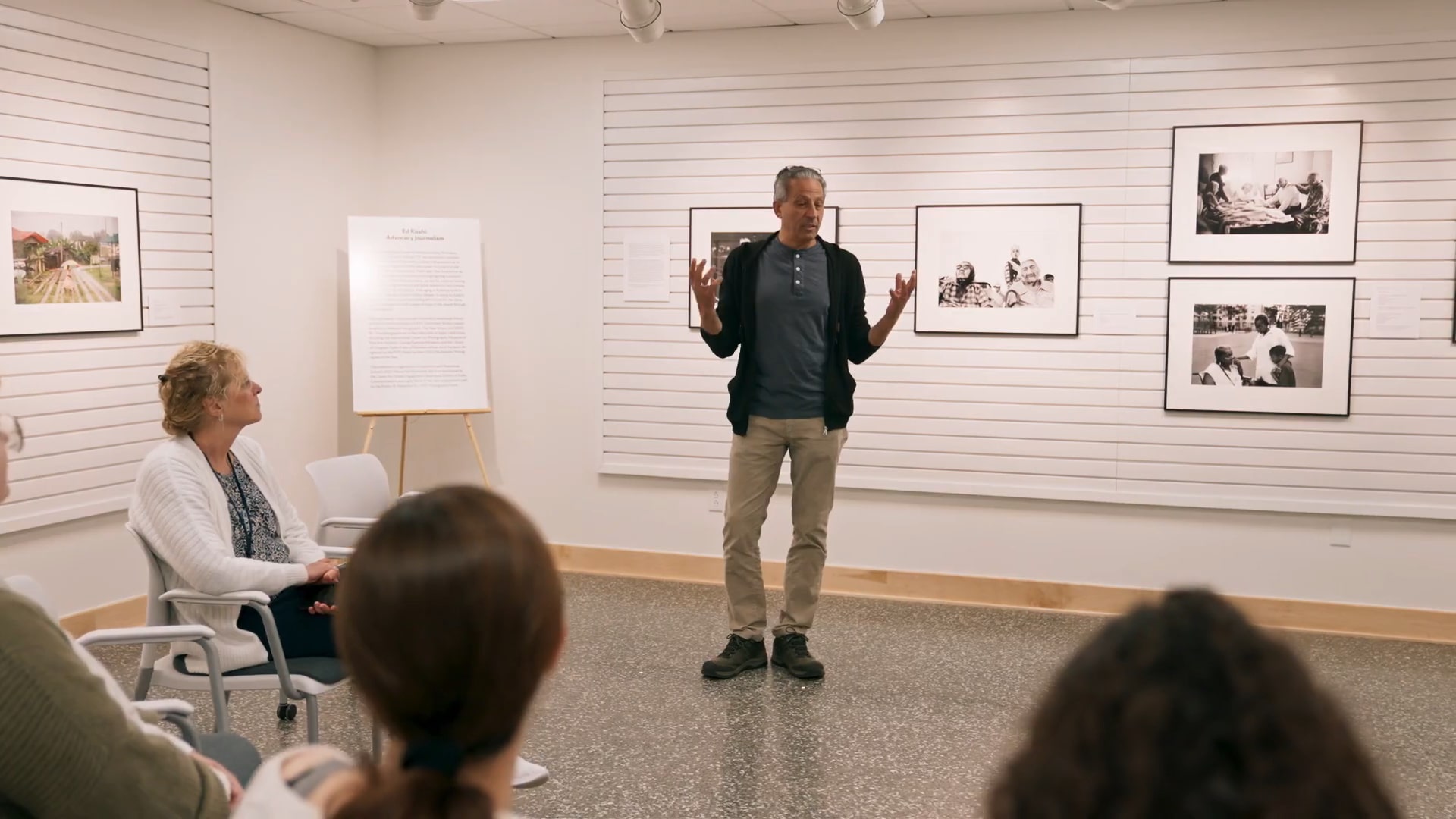 A speaker presenting to a group in a gallery-like setting with framed black-and-white photographs on the walls