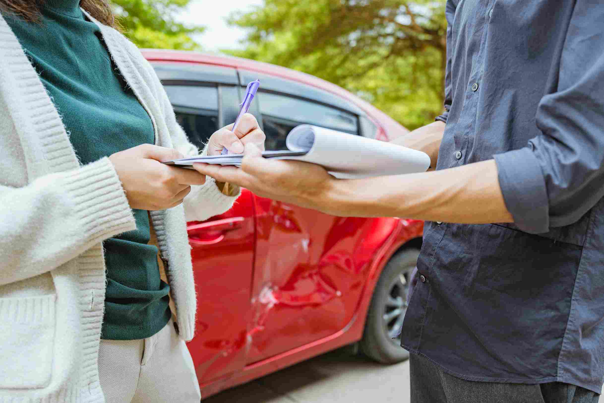 Two people exchanging information and writing notes after a car accident beside a damaged vehicle