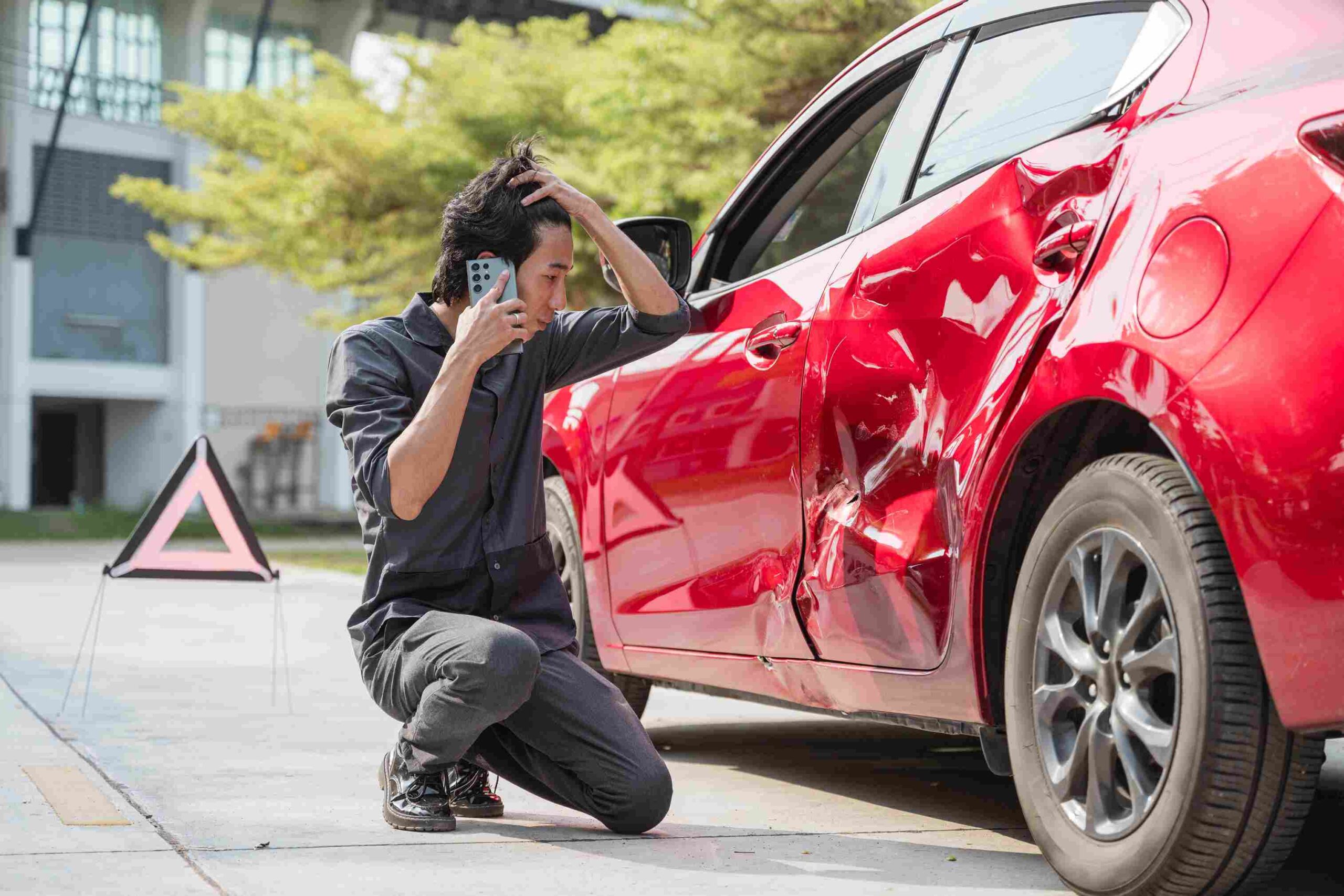 A man looking stressed while inspecting damage on a red car after an accident and talking on the phone