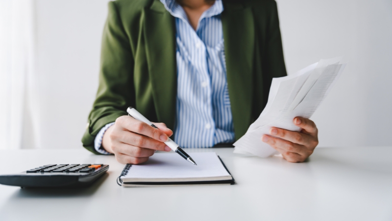 Person reviewing bills and writing notes next to a calculator while analyzing personal debt and expenses
