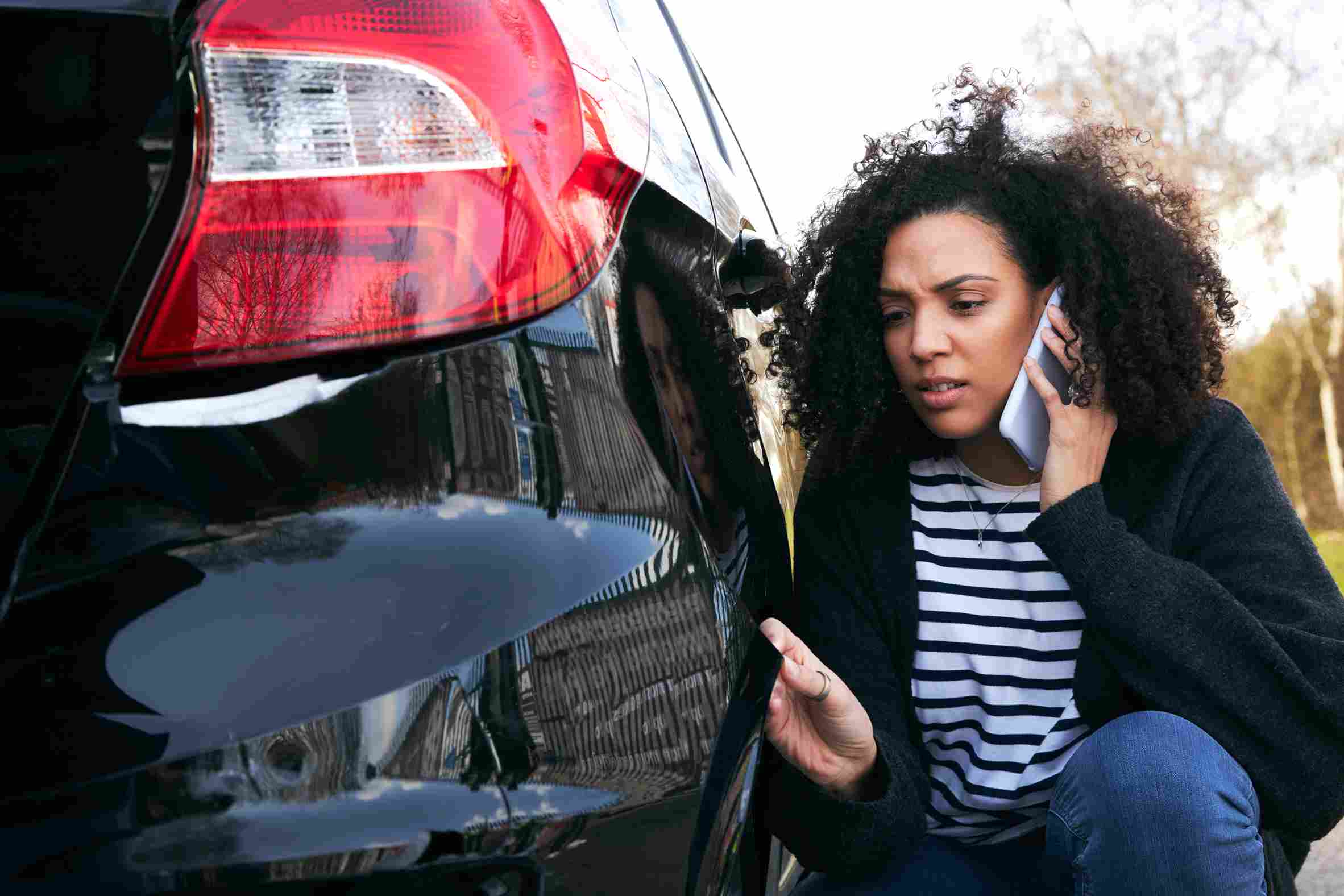 A woman inspecting damage on her car while talking on the phone after a hit and run accident