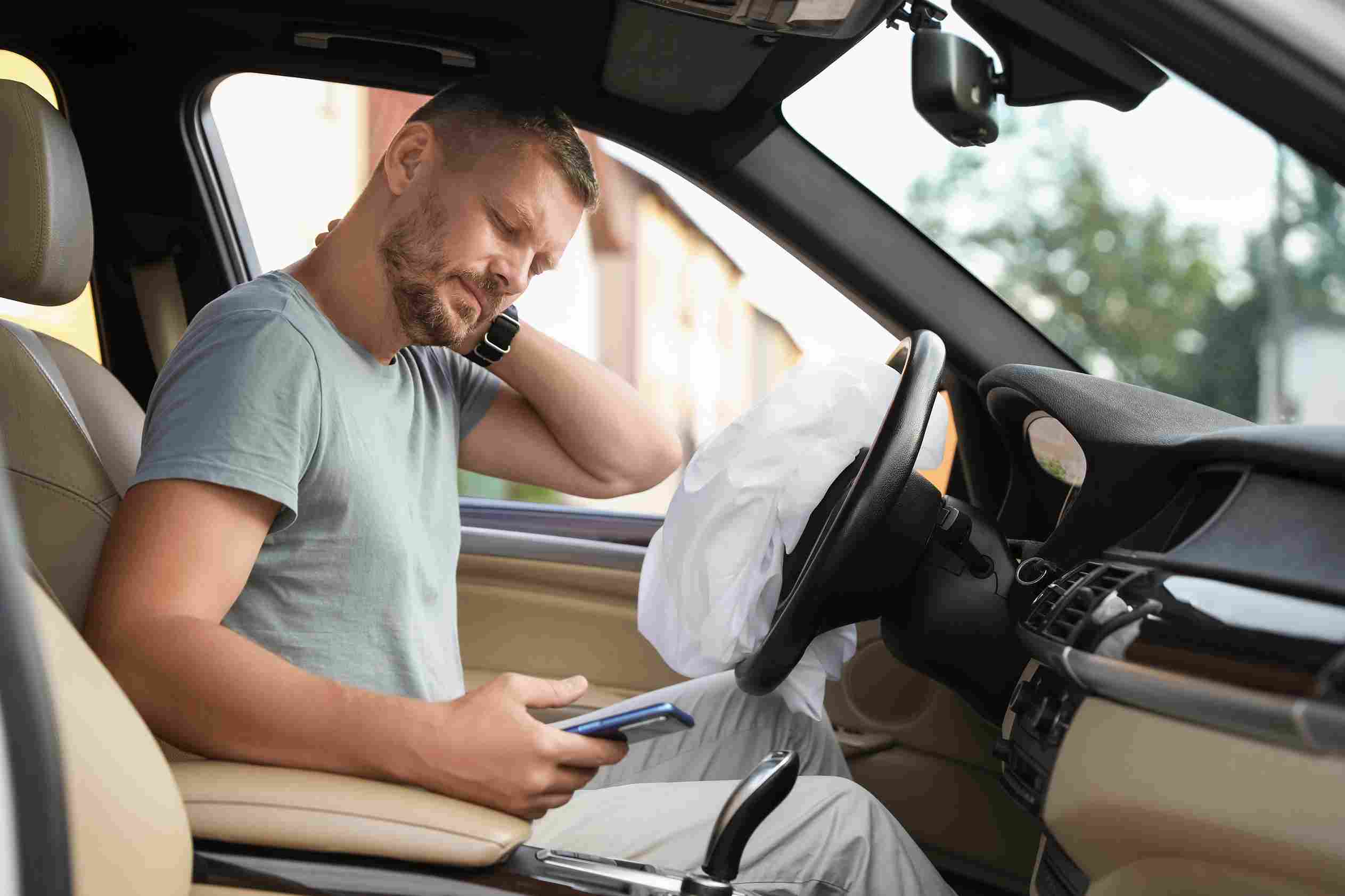 A driver sitting in a car after an accident holding his neck while looking at his phone with a deployed airbag visible