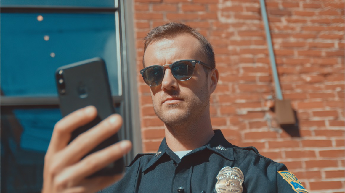 Police officer outdoors looking at a smartphone while standing in front of a brick building