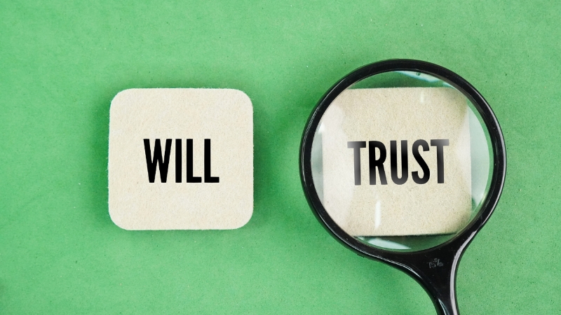 Cards labeled “Will” and “Trust” on a table with a magnifying glass, illustrating why estate plans often include both
